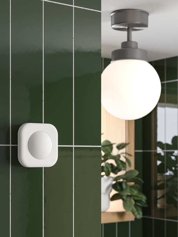 A VALLHORN wireless motion sensor is placed on a bathroom wall close to a FRIHULT ceiling lamp in stainless steel color.