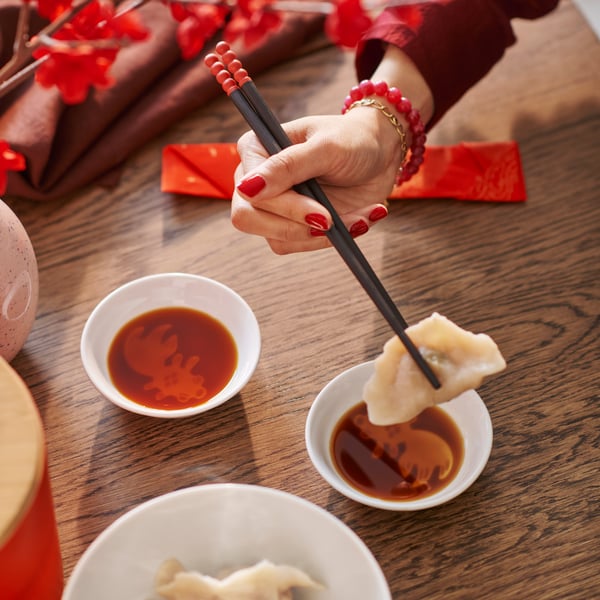 A hand holding chopsticks lifts a dumpling from a FÖSSTA sauce dish filled with dark sauce.