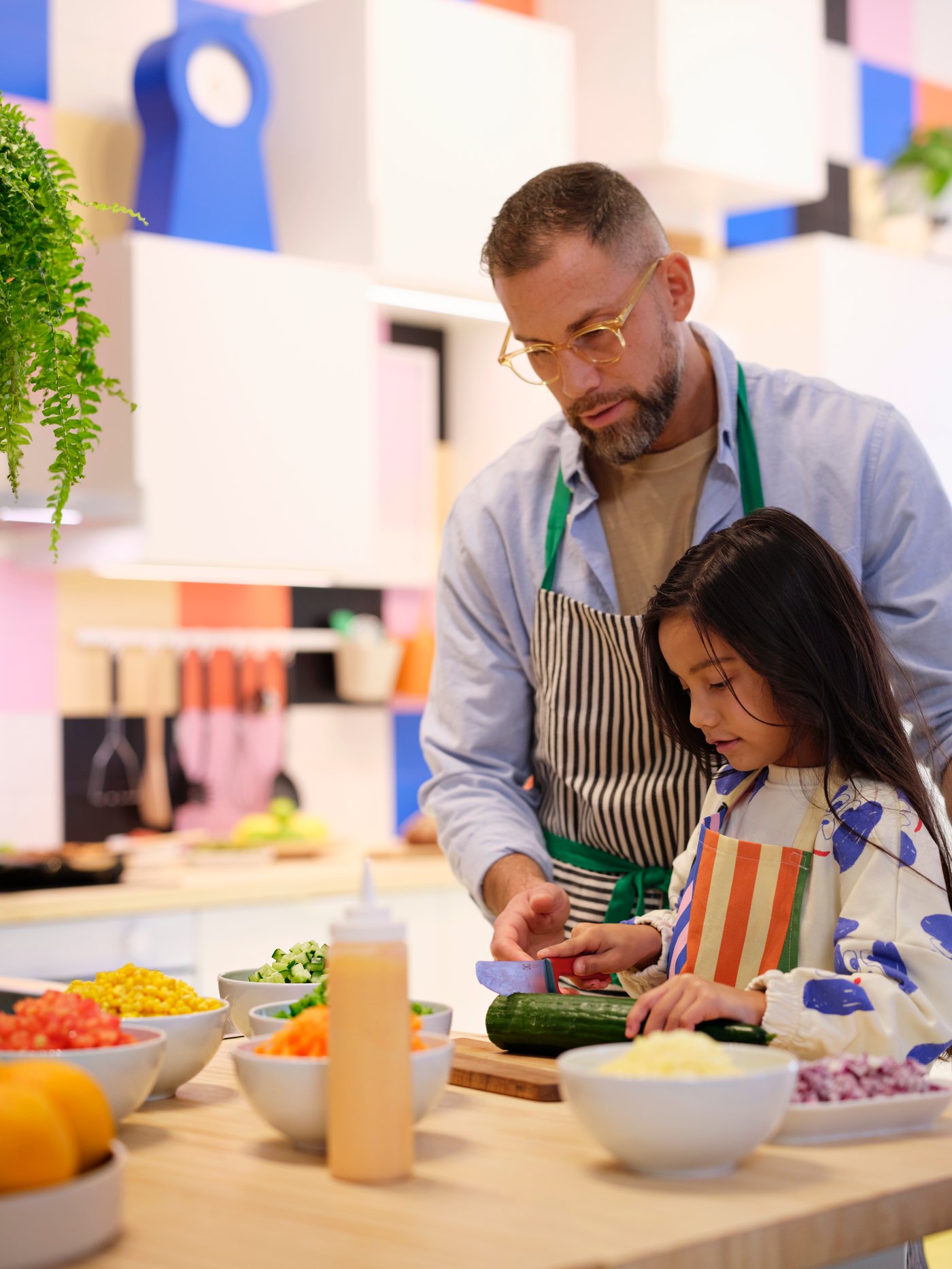 A grown-up is helping a child cut vegetables in a kitchen. There are many white IKEA 365+ bowls with veggies on the worktop.
