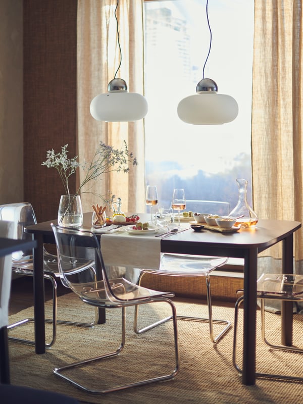 A STRANDTORP extendable table in front of a large window, laid for a meal for two with flowers, food dinnerware and glasses.