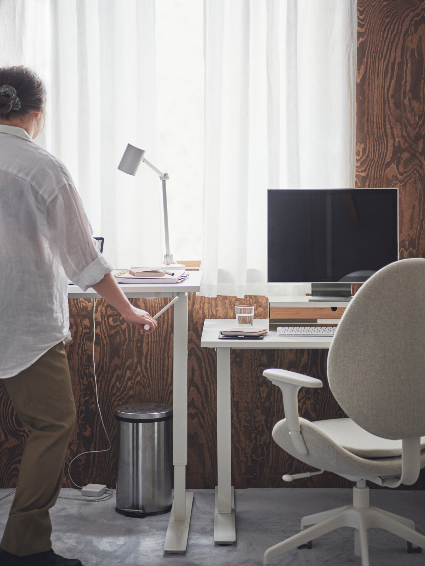 A person standing by a white TROTTEN desk holding its height-adjusting crank handle. Next to it is an identical, lower desk.