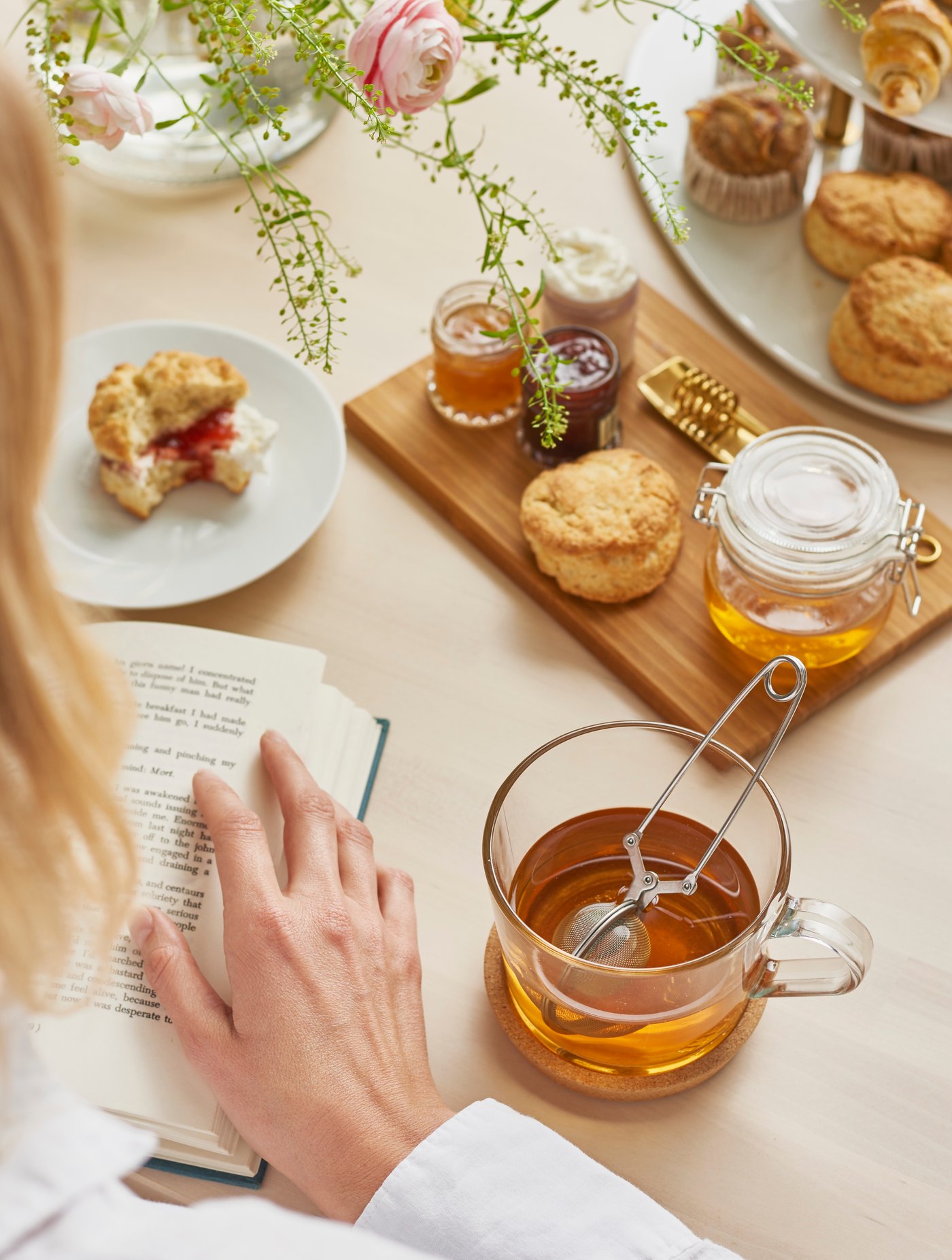 Person liest ein Buch, daneben stehen eine Glas-Teetasse mit Teesieb, Honigglas, kleine Marmeladen und frische Gebäckstücke auf einem hellen Tisch – eine ruhige Pause mit Tee und Süßem.