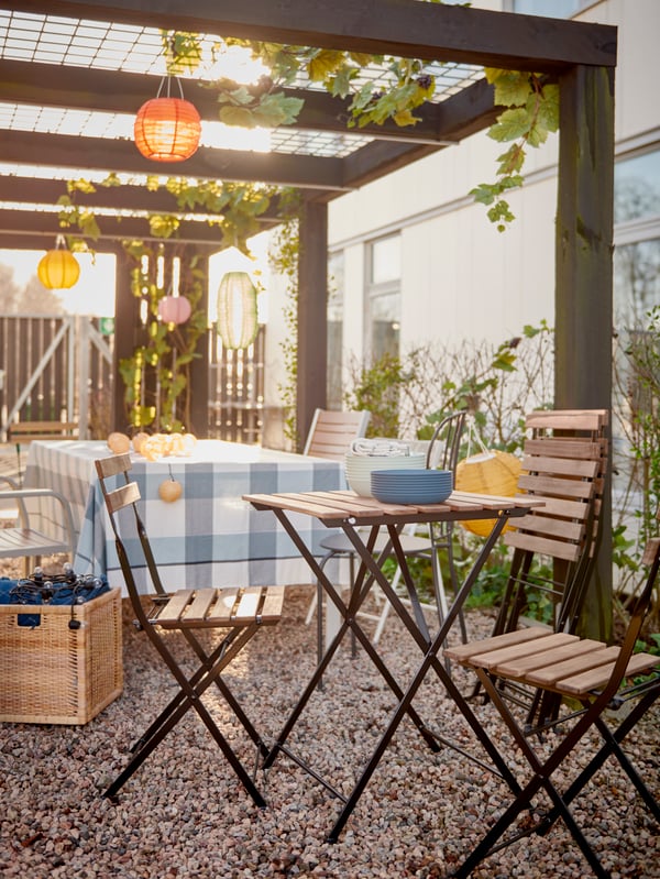 Una mesa plegable TÄRNÖ junto a cuatro sillas, dos armadas y dos plegadas, en un patio con suelo de gravilla. Detrás del pequeño comedor de terraza se ve una mesa más grande cubierta por un mantel a cuadros celestes y sillas metálicas con apoyabrazos bajo un parrón de madera.