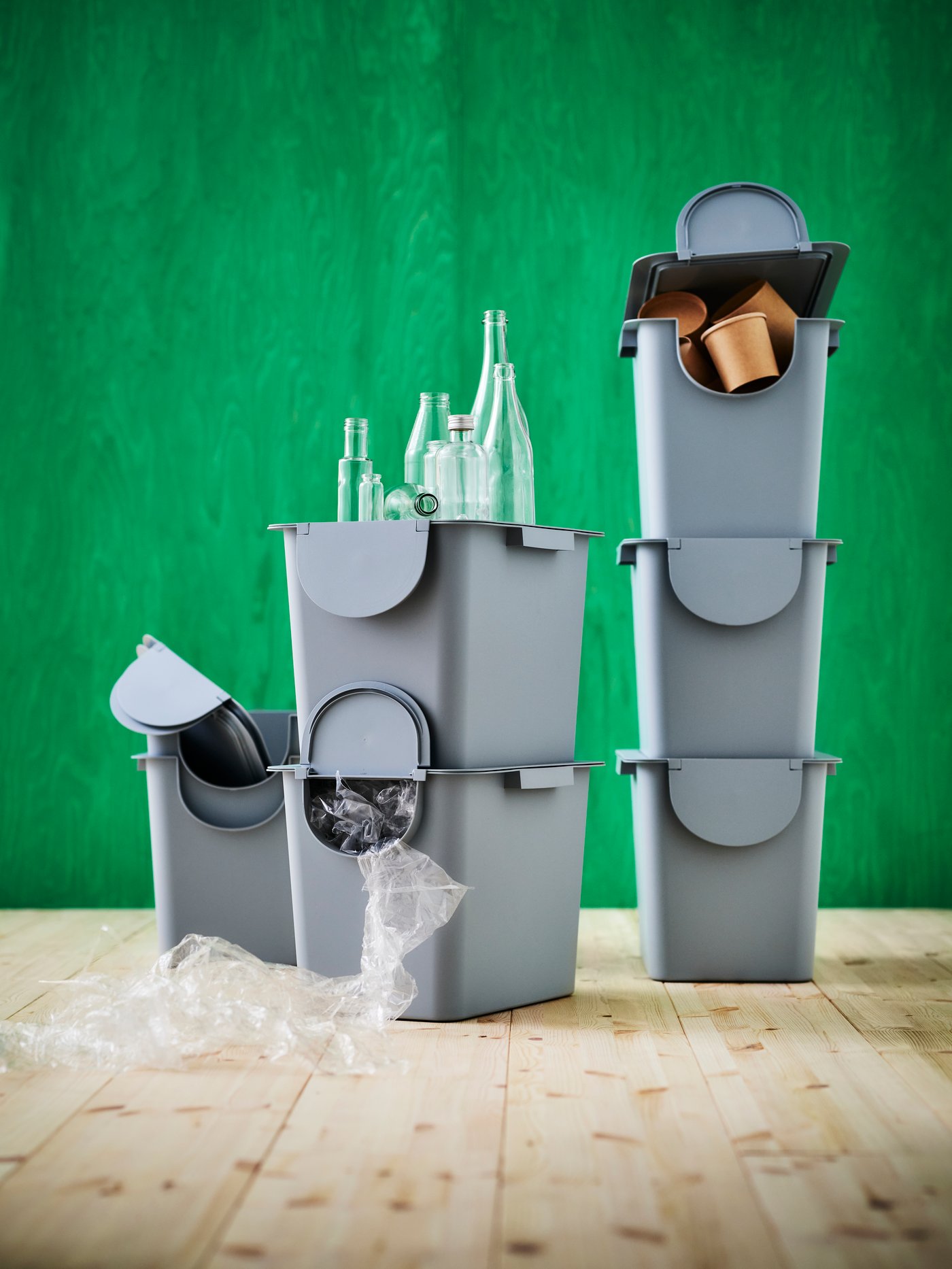 Light grey-blue TRÖSKNING bins with lid stand stacked on top of each other on a wooden floor against a green background.