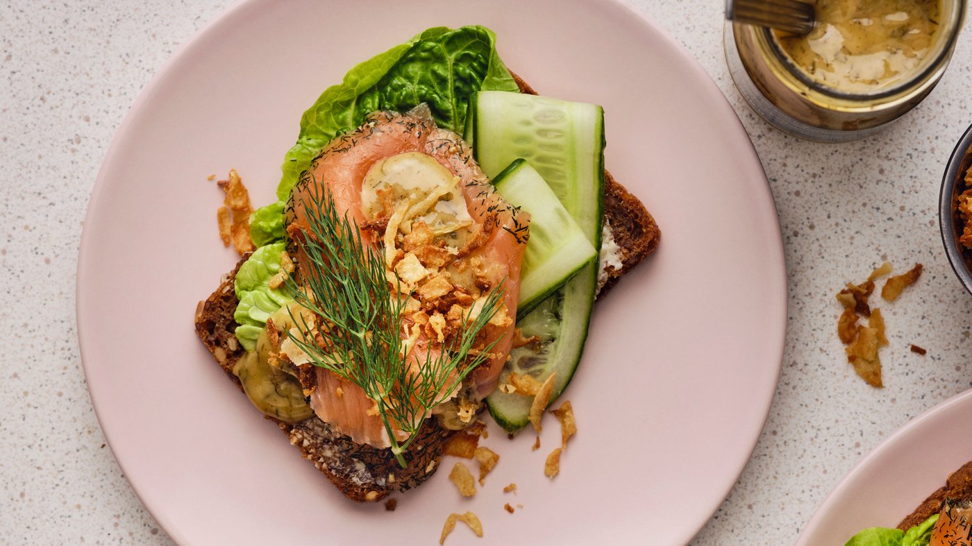 Una fetta di pane rustico scuro con diverse fette di salmone, lattuga, cetriolo, aneto e una salsa alla senape su un piatto rosa.