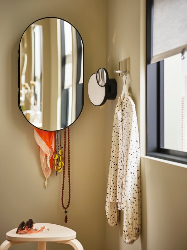 Miroir LINDBYN noir avec rangement pour divers accessoires, à côté de trois crochets muraux avec un chemisier sur un cintre blanc.