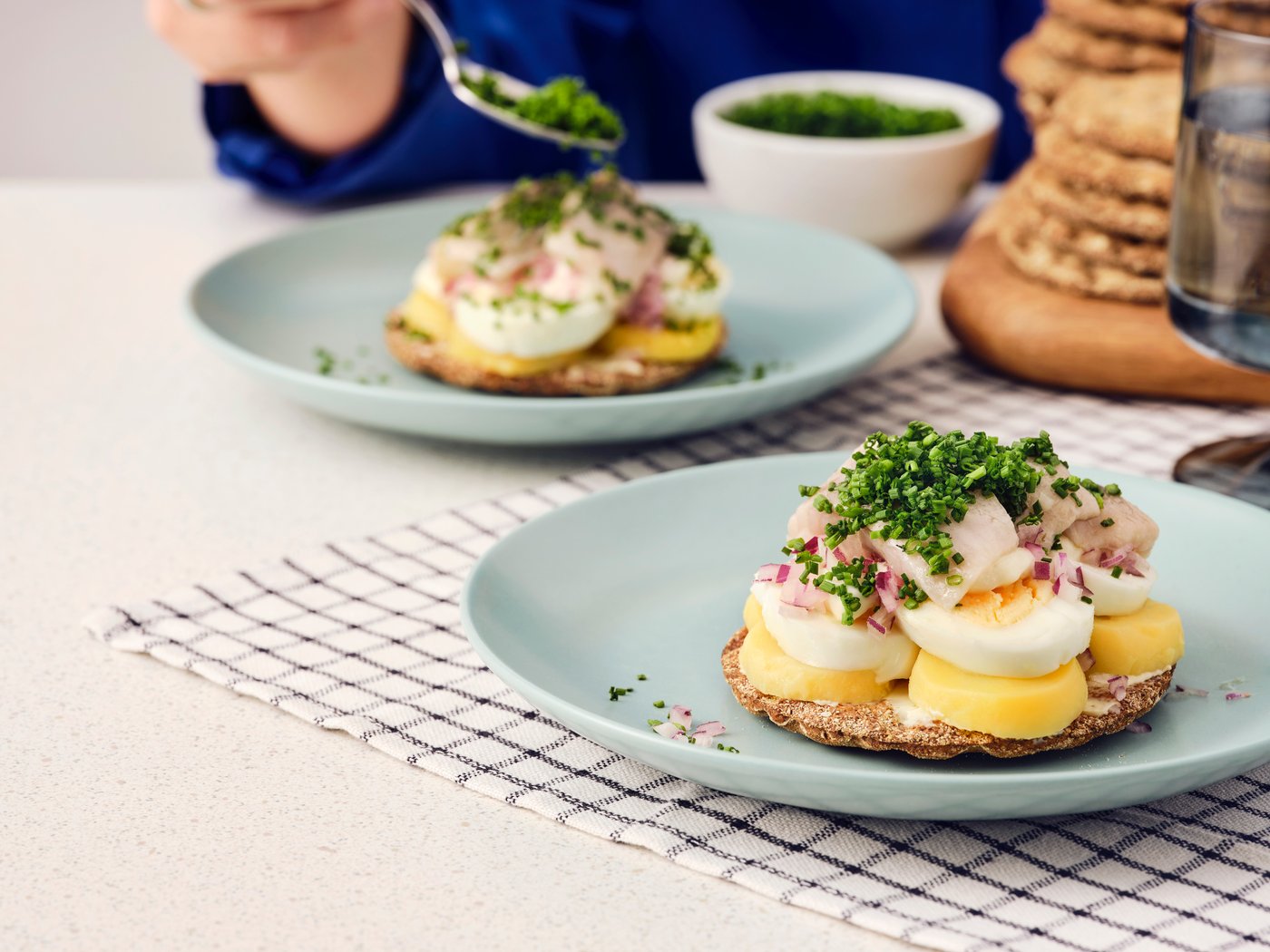 Två öppna mackor med KNÄCKEBRÖD RÅG rågknäckebröd, ägg, SILL DILL marinerad sill och gräslök på blå tallrikar.