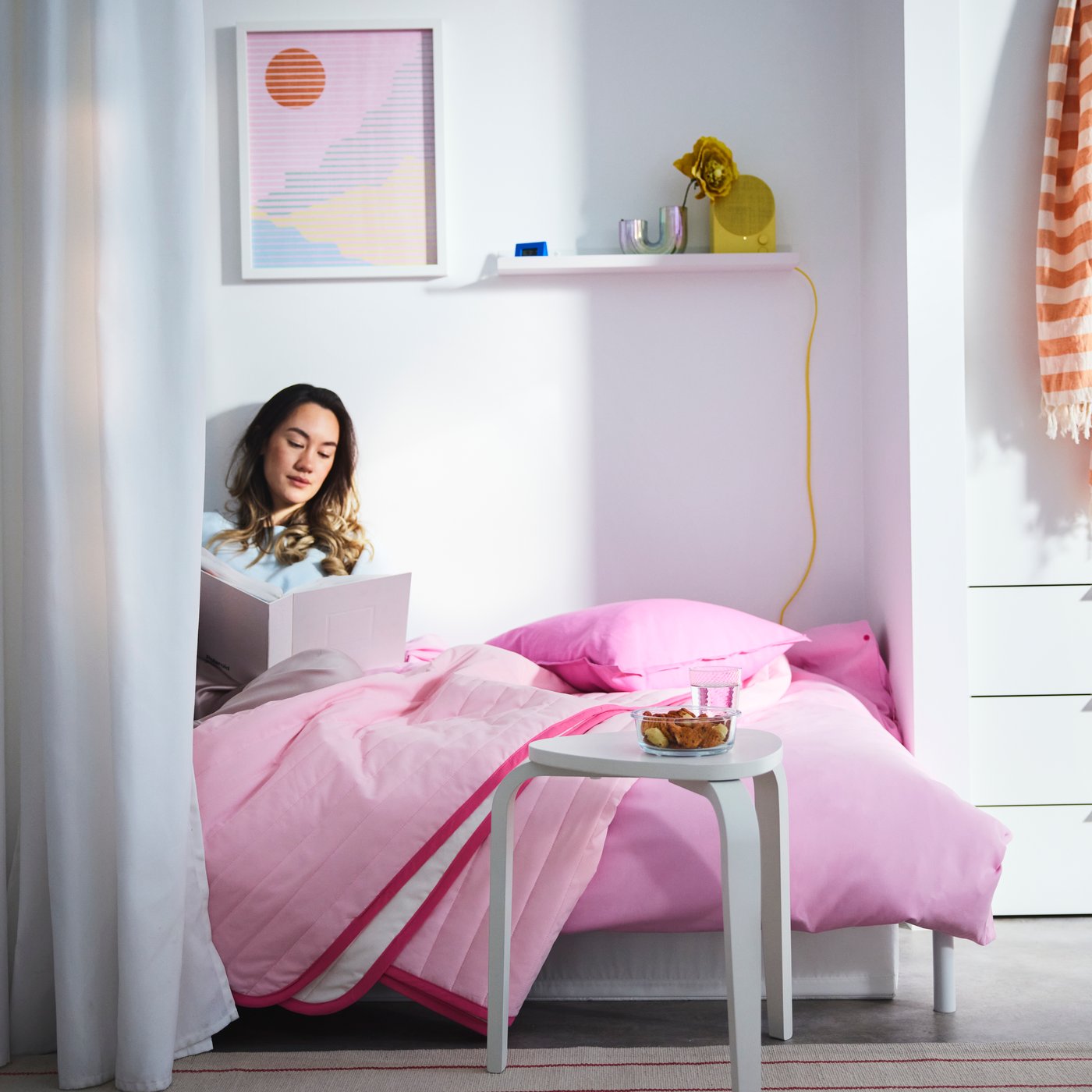 A person sitting studying in a bed made with the pink VETEJORDFLY bed spread, placed in a white room with pops of colour.