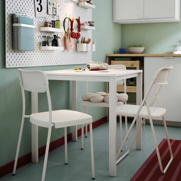 A white VIHALS table is set with green dishes, surrounded by a chair, with organised wall shelves in a light room.