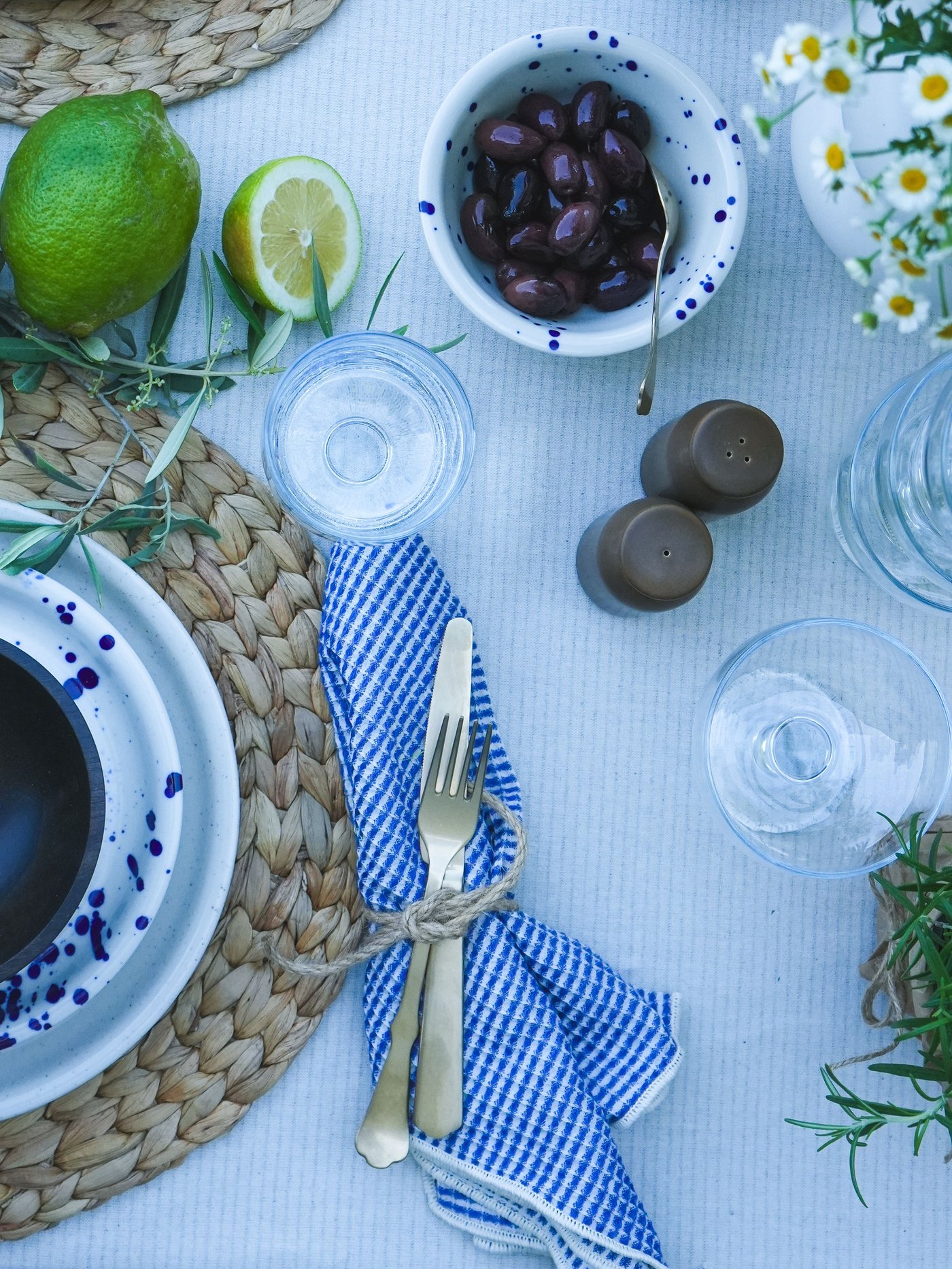 Gedeckter Tisch mit rustikalem Besteck, blau-weissem VÄLVÅRDAD Küchenhandtuch, einer Schale voller Oliven, frischen Zitronen und floralen Verzierungen.