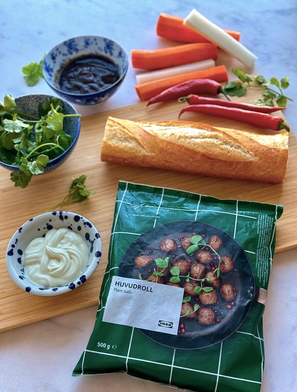 Eine Auswahl an Zutaten für eine Mahlzeit: frische Kräuter, Saucen, Chilischoten, Karotten, ein Baguette und ein Fertiggericht auf einer Holzunterlage.