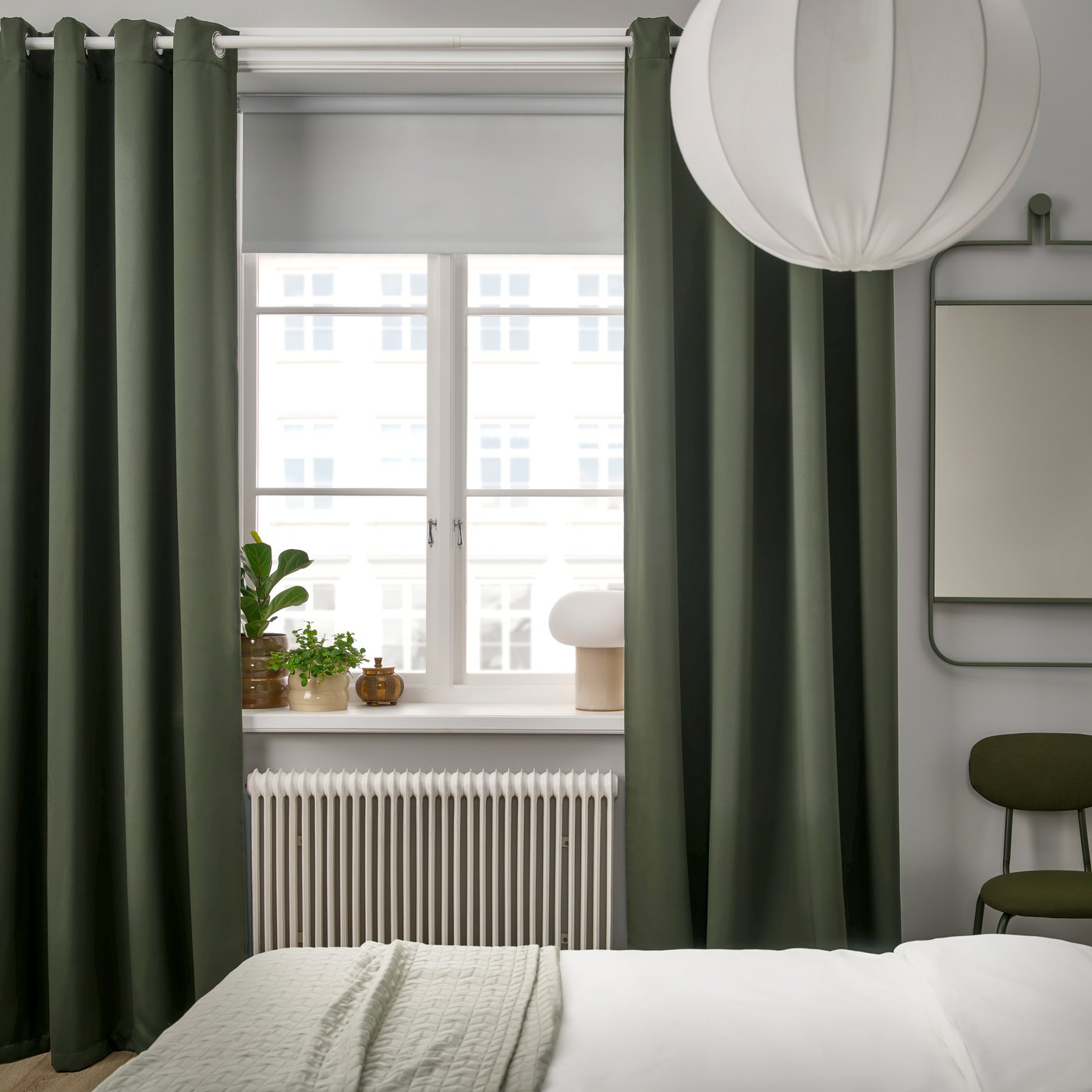 Dark green MAJGULL blackout curtains with eyelet heading hang floor-to-ceiling in front of the window, effectively darkening the room. The heavy fabric provides privacy, reduces incoming light, and at the same time adds a decorative touch to the bedroom.