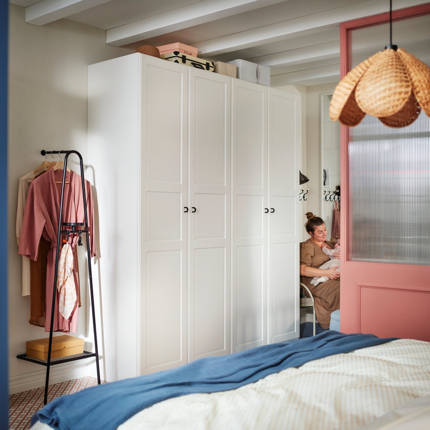 A woman and baby sit at the end of a white PAX/TYSSEDAL wardrobe with a black ÄLGANÄS clothes rack with shoe storage by it.