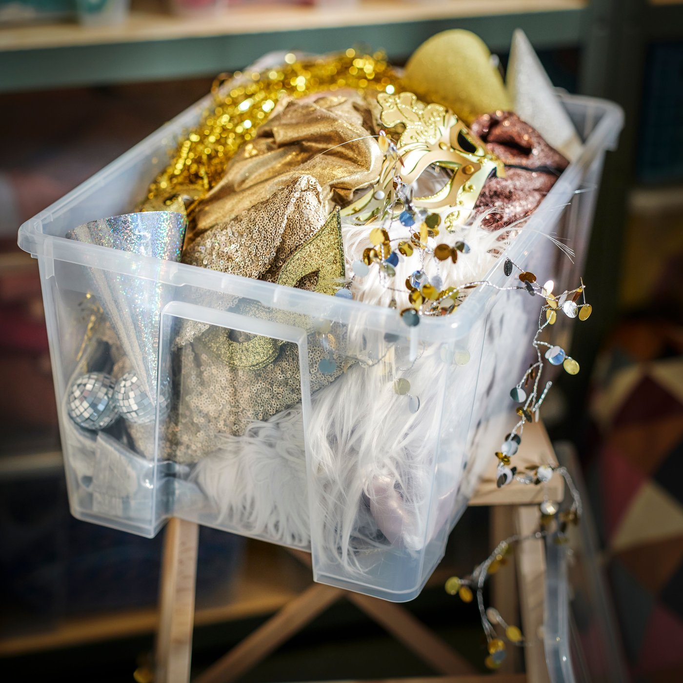 A transparent SAMLA box with a lid filled with glitter clothing and pieces of fabric.