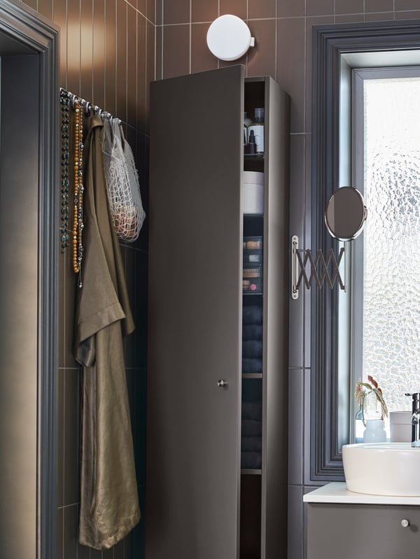 A dark-grey high cabinet, a round mirror with an extendable arm and a dark-grey/white wash-stand.