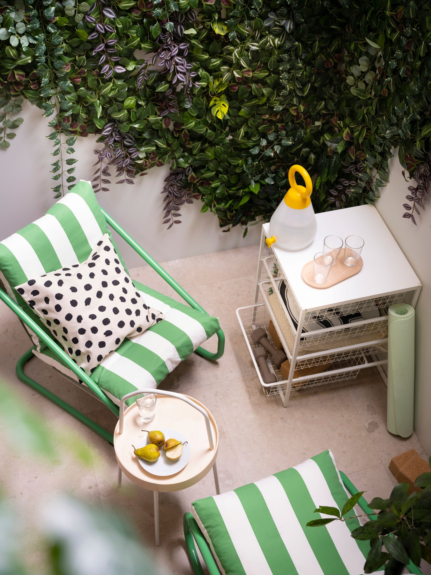 A cozy corner featuring a green and white striped lounge chair with a polka-dotted pillow, next to a white side table holding a modern lamp, set against a lush wall of greenery.