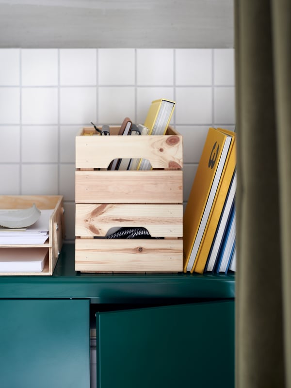 Dos cajas de madera KNAGGLIG apiladas con libros, dos bandejas de madera para cartas y algunos libros encima de un gabinete verde.
