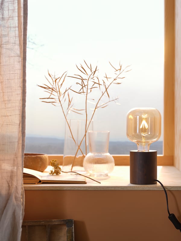 Vases transparents et lampe de table avec pied noir et ampoule décorative sur un appui de fenêtre avec rideaux/voilages beiges