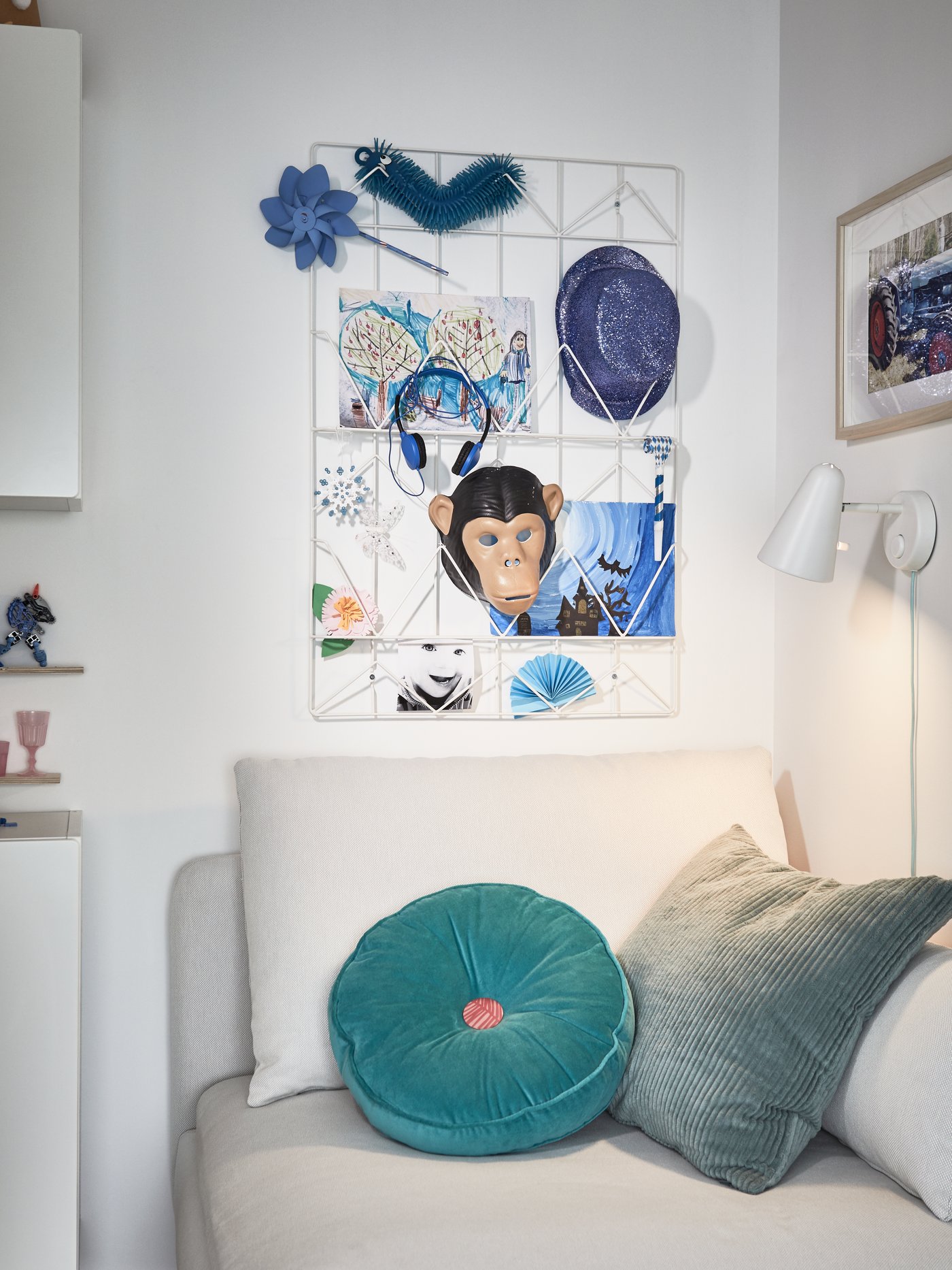 A white wired memo board with blue drawings and accessories, a beige sofa module, torquoise cushions, a white wall lamp.