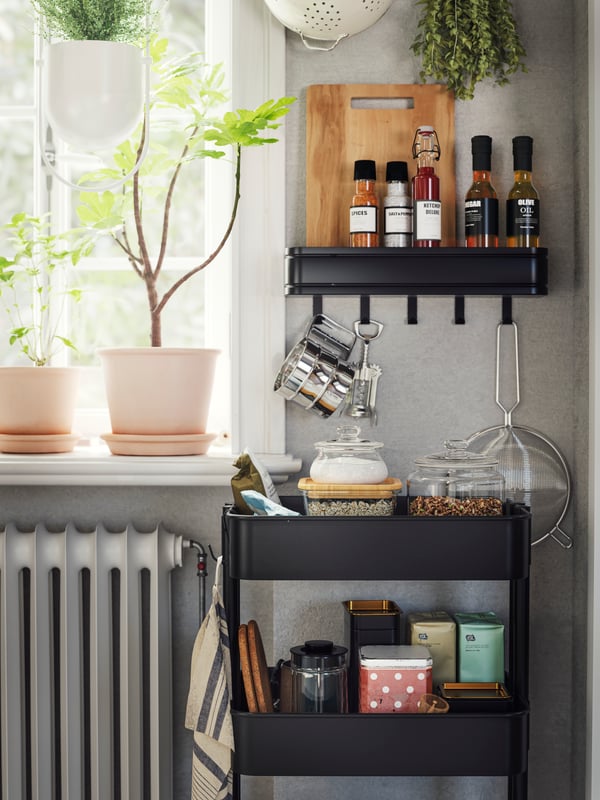 A black RÅSKOG trolley next to a window has food containers and tins stacked on it and a black shelf with hooks above it.