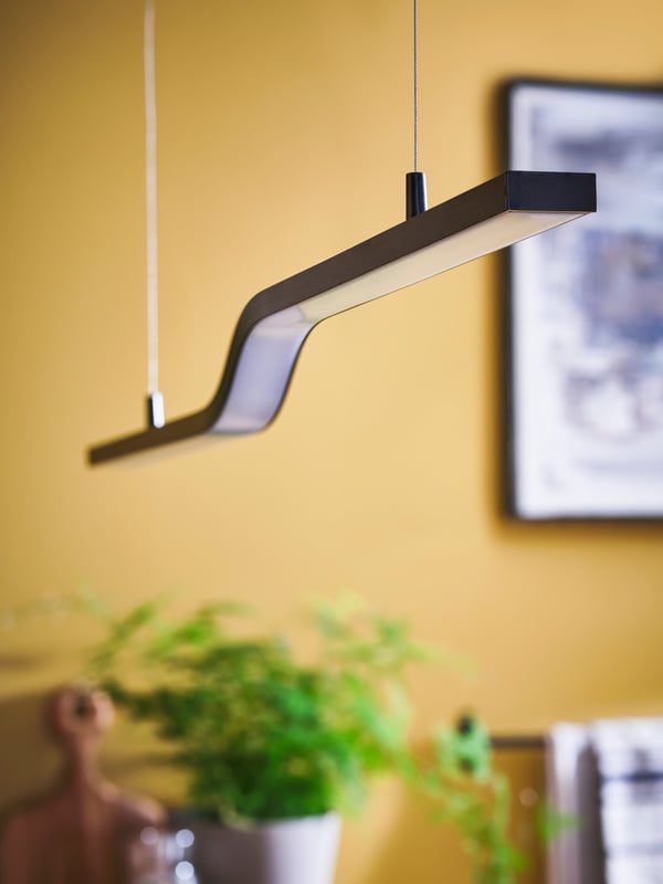 A black PILSKOTT LED pendant lamp hanging in the middle of a yellow room with a picture in a black frame on the wall.