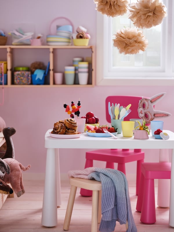 Stanza in tinta pastello con seggioline MAMMUT rosa intorno a un tavolo per bambini MAMMUT bianco, con snack colorati.