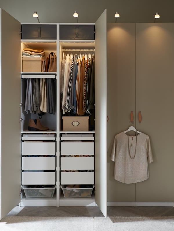 Side-by-side PAX wardrobes with grey-beige REINSVOLL doors. One wardrobe is open, revealing neatly organised clothes inside.