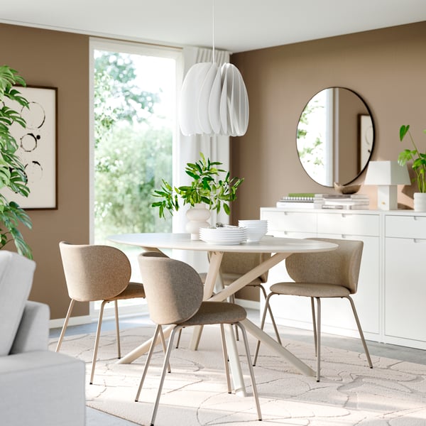 Een lichte IKEA-eetkamer met een ronde witte tafel, vier beige stoelen, een sculpturale hanglamp, een wit dressoir met ronde spiegel en weelderige groene planten baadt in natuurlijk daglicht.