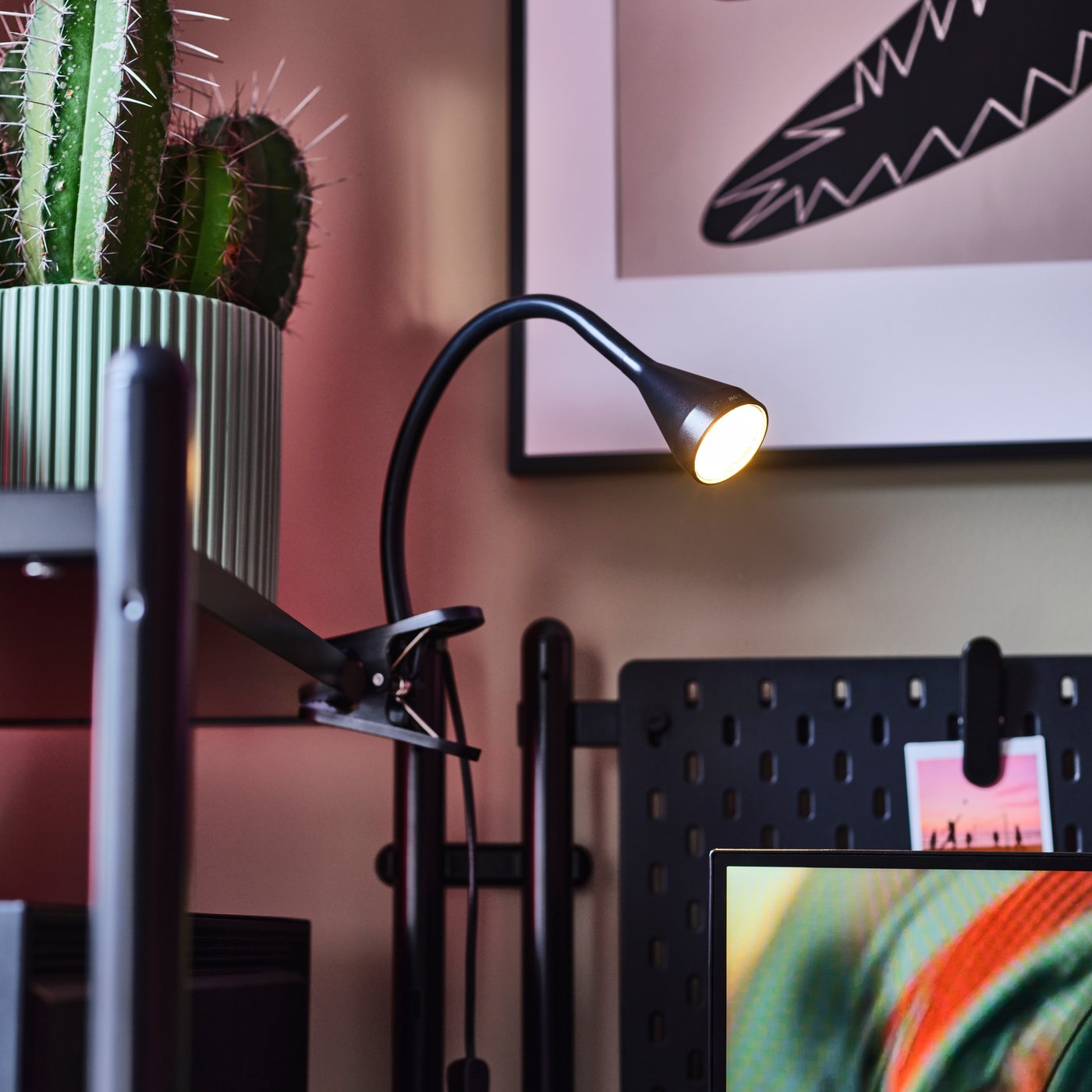 Un espacio de trabajo con un foco LED de pinza NÄVLINGE negro iluminando un escritorio junto a un cactus verde brillante en maceta.
