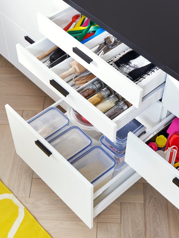A kitchen with open white drawers displaying plastic food containers, kitchen utensils and kitchen tools inside.