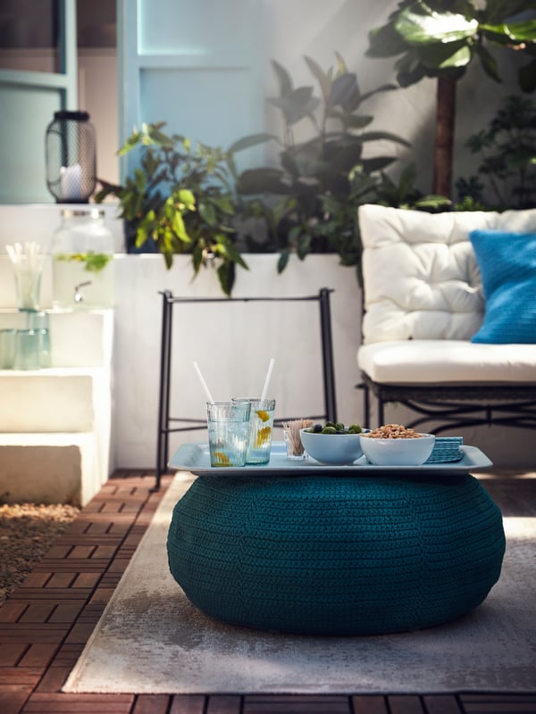 A blue tray with bowls of snacks and glasses of lemonade on dark green pouffe, with its table legs in the background.