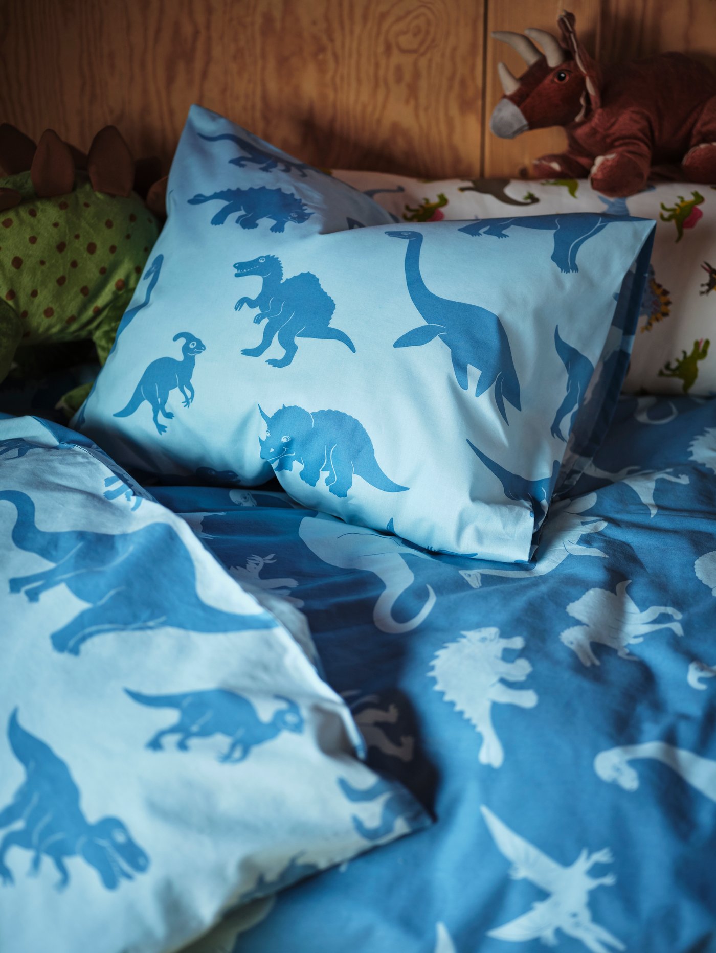 A child’s bed with a light blue JÄTTELIK quilt cover and pillowcase with dinosaur prints and two JÄTTELIK dinosaur soft toys.