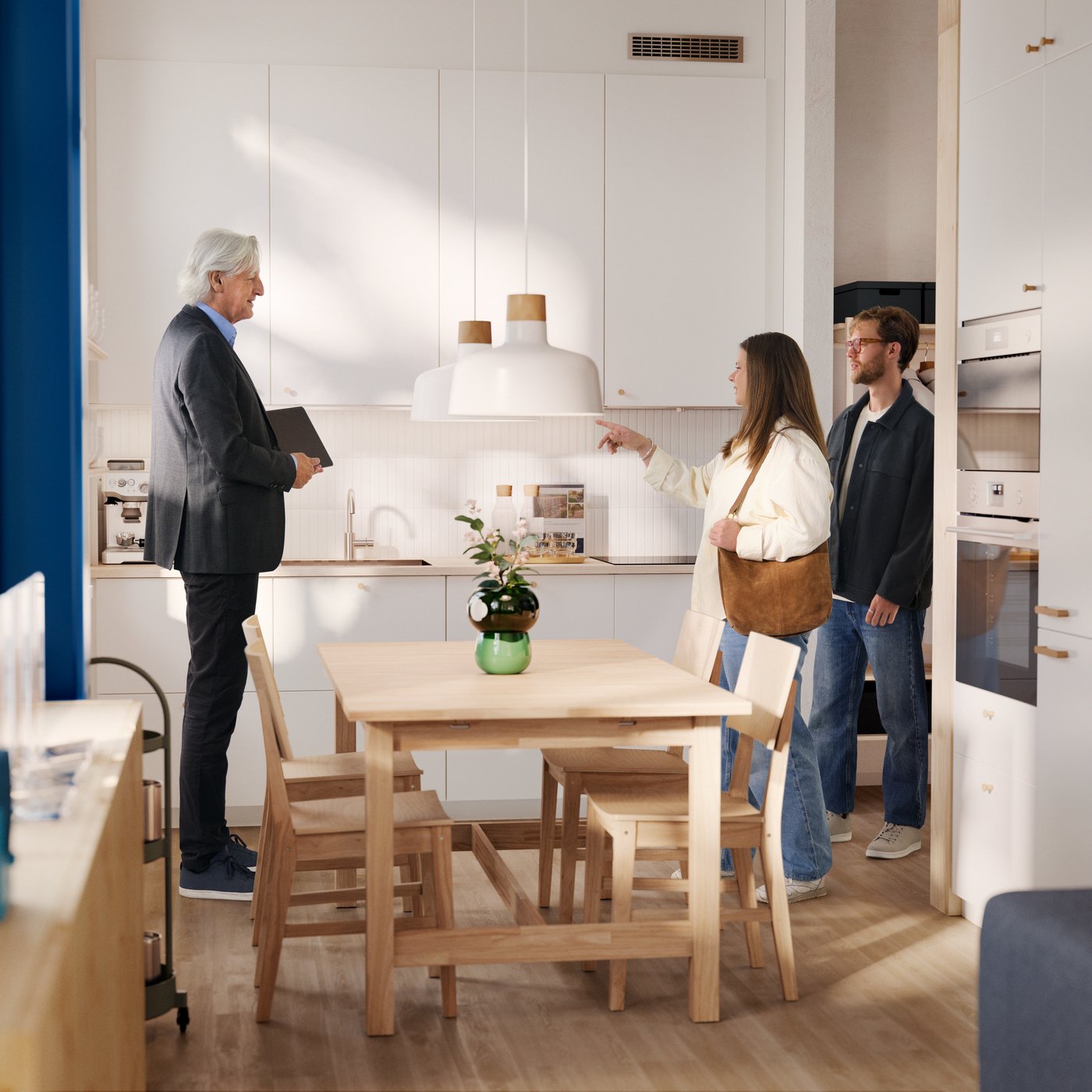 An estate agent in a suit greets a casually dressed young couple in a small modern kitchen with white cabinets and pine table.
