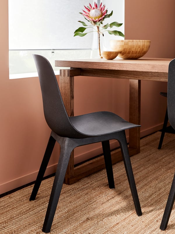 A black ODGER chair on a jute rug by a window with blind, table in front with two wooden bowls and a vase with a flower.