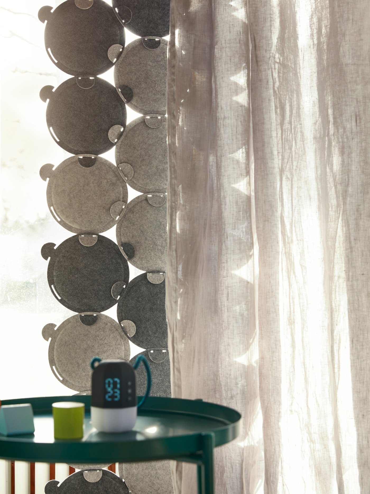 ODDLAUG sound absorbing panels hanging in a window with neutral-colored curtains in front of it.