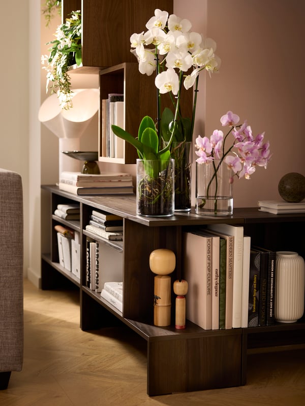 HOLMERUD side tables filled with books, decoration and plants follow the outline of an outward corner in a sunlit room.