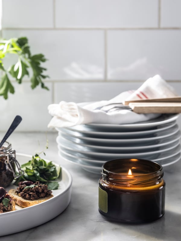 A lit PRAKTRÖNN scented candle in glass with lid by canapés and tableware on a marble worktop with a white-tile splashback.