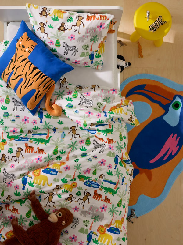 Birds-eye view of white children’s bed with colourful jungle-themed bedding, cushion with tiger motif and toucan rug.