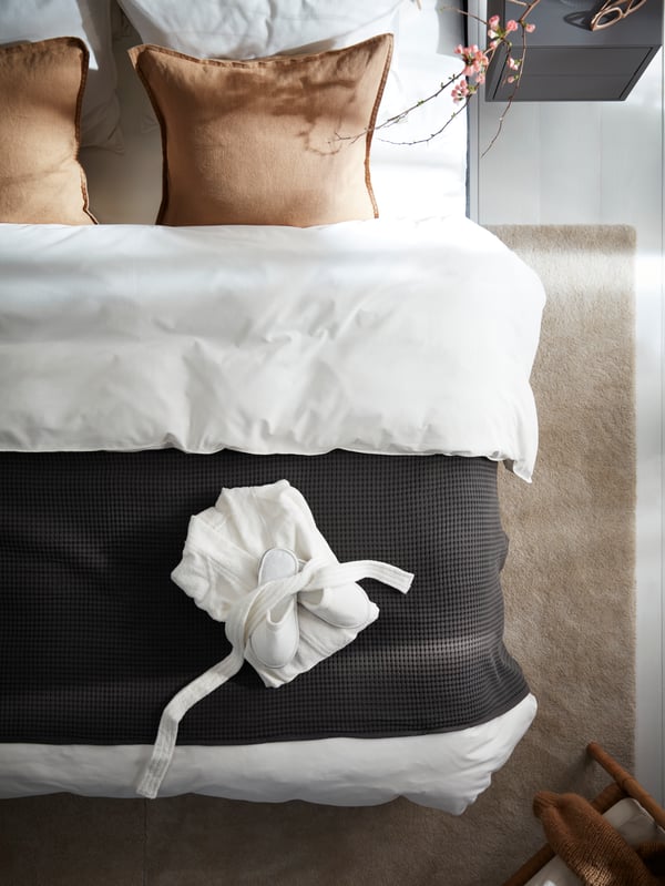 A bed with white ÄNGSLILJA bed linen, a dark grey bedspread and a white bath robe with slippers, on an off-white STOENSE rug.