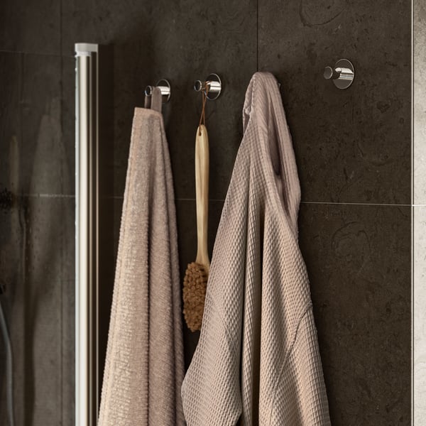 Un albornoz BJÄLVEN beige con textura en cuadrícula colgado en la pared junto a una toalla VALLASÅN beige y un cepillo corporal de madera.