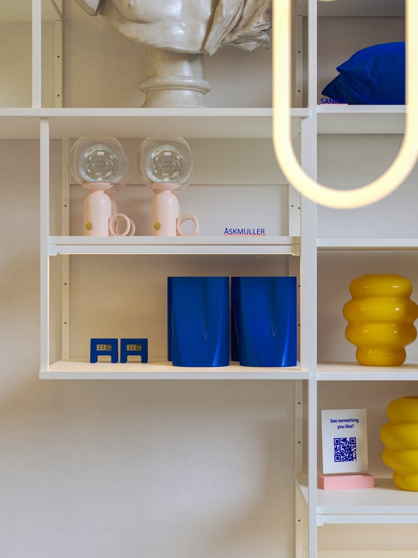 Scaffali a giorno PLATSA bianchi montati a parete su cui sono esposti oggetti decorativi per la casa in giallo, blu e rosa pallido e un cartellino con codice QR. 