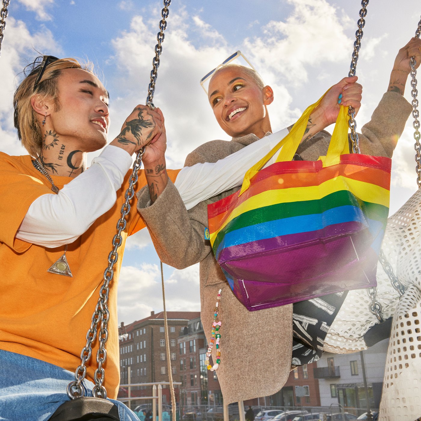 Twee mensen zitten op schommels buiten met een IKEA draagzak in regenboogkleuren die tussen hen in hangt. De twee mensen zitten in een stadse omgeving met gebouwen en een mooie hemel.