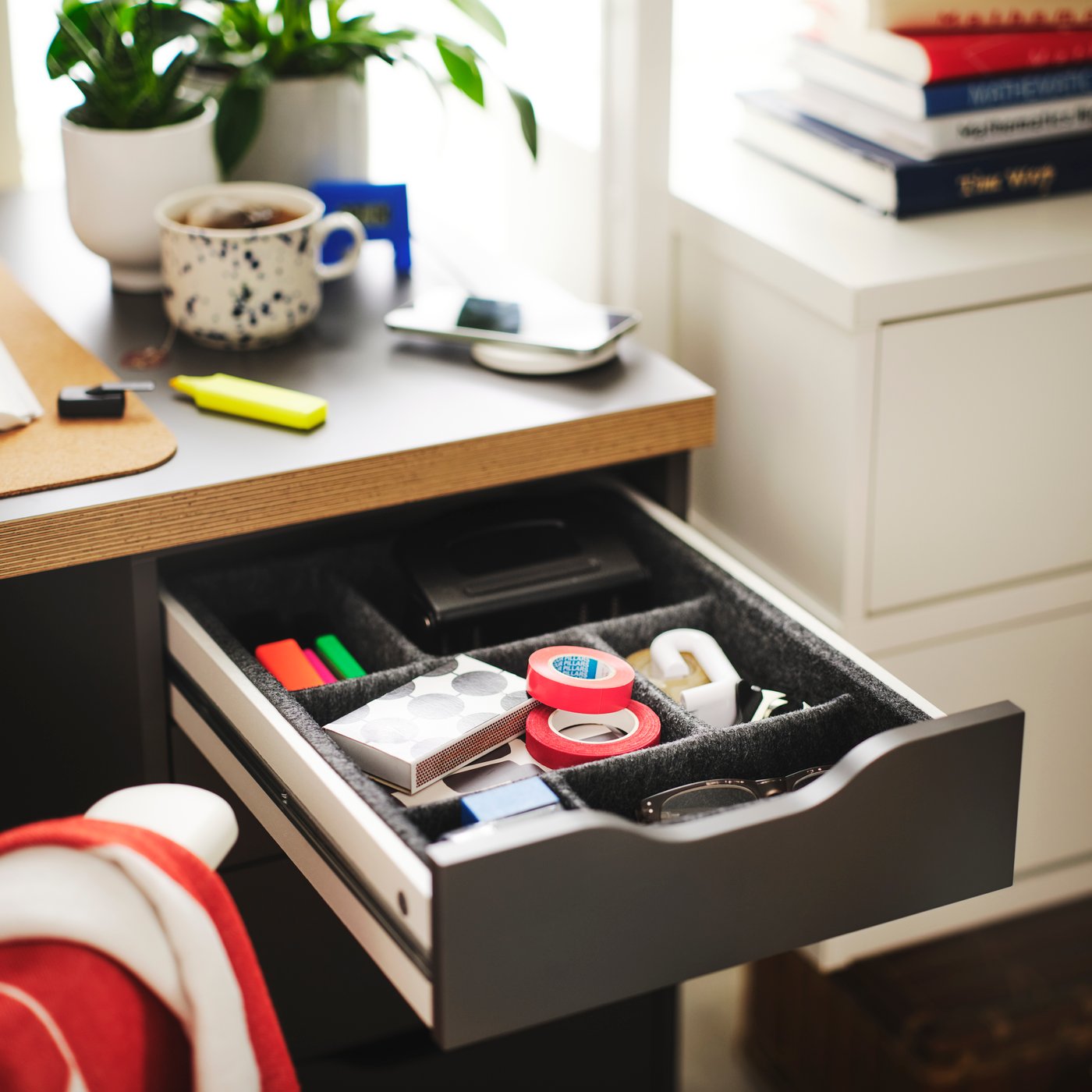 A dark grey ALEX drawer unit with neatly organised supplies, a plant, and a coffee cup on a desk.