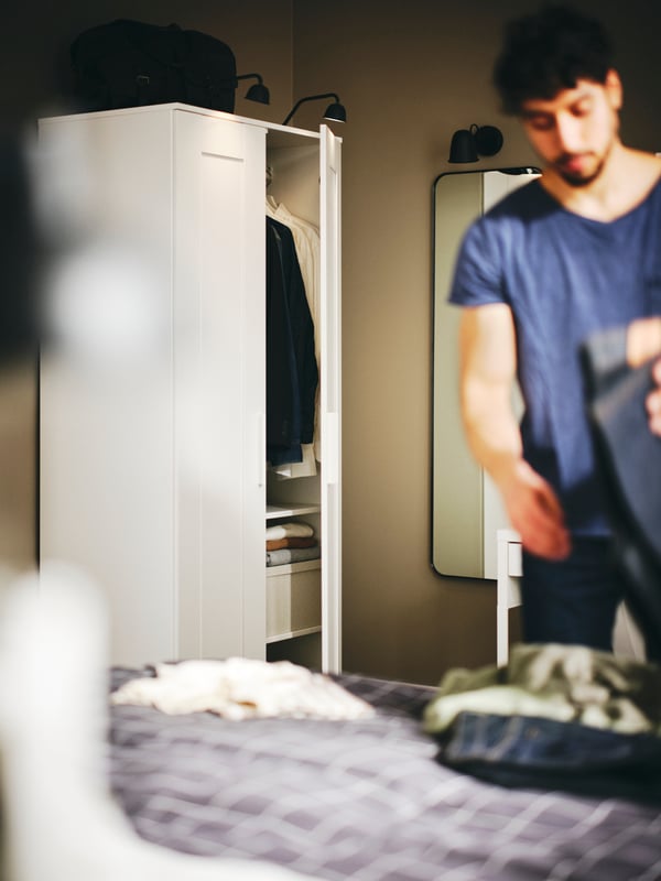 A person folding clothes on a bed with a black LINDBYN mirror and a white BRIMNES wardrobe with 2 doors behind them.