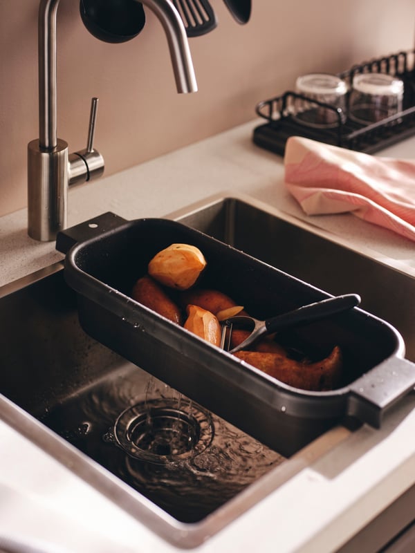 IKEA LILLHAVET colander in black resting on the edge of a stainless steel kitchen sink on a white worktop