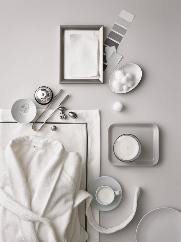 A white ROCKÅN bathrobe, white/dark grey bed linen, a small silver frame, white bowls and cups with sugar lumps and candles.