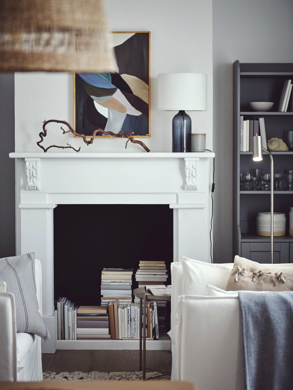 Dans un séjour lumineux, des livres s’empilent dans la cheminée déco, près d’une armoire HAUGA grise et d’un fauteuil BACKSÄLEN blanc.