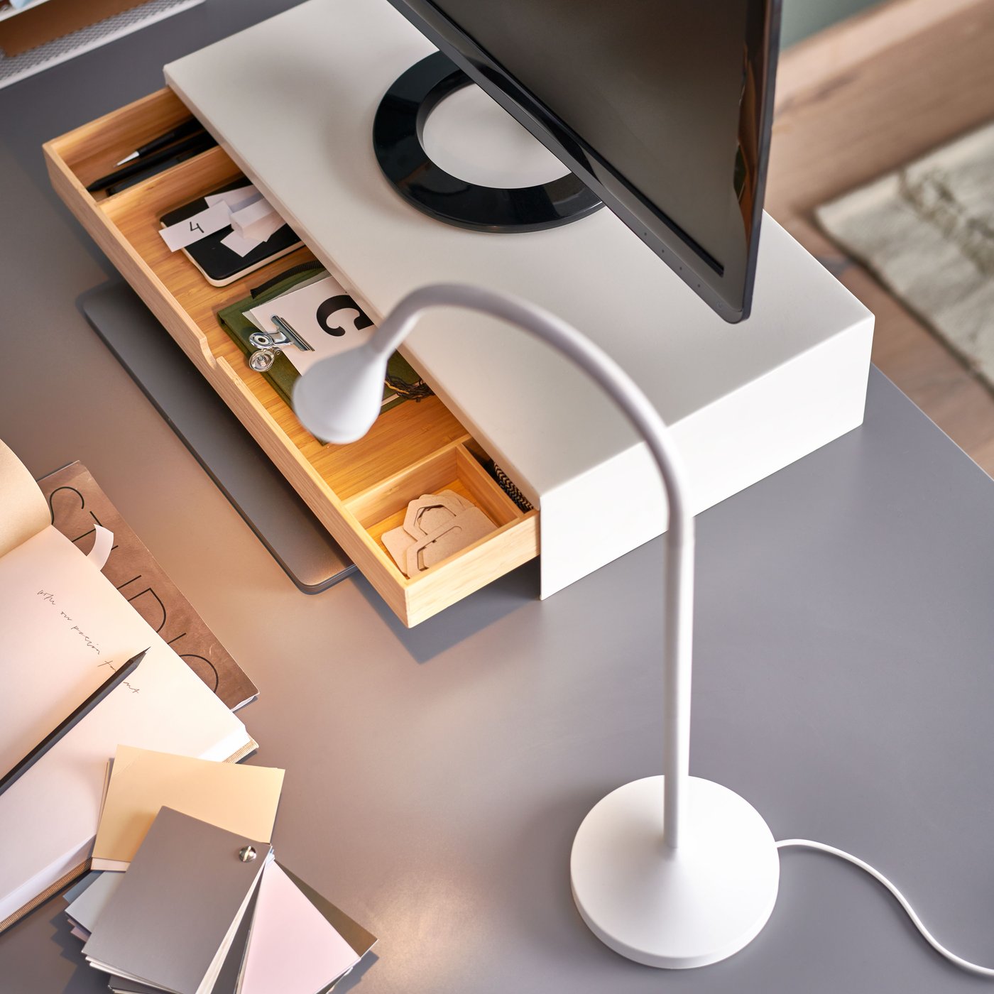 A white NÄVLINGE LED work lamp on a grey table top, beside an ELLOVEN monitor stand with a drawer that’s open.