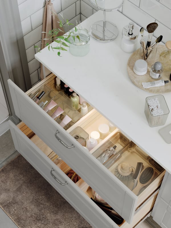 A wash-stand with toiletries on top and two open drawers with GODMORGON boxes with compartments inside containing make-up.