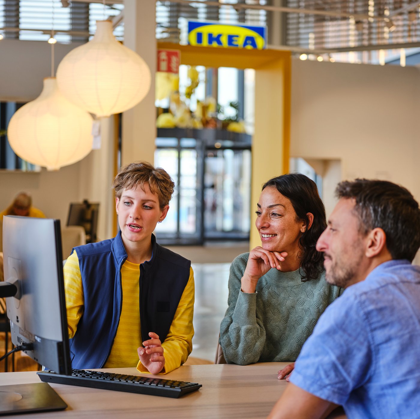 Drei Personen an einem Beratungstisch in einem IKEA-Einrichtungshaus; links eine Mitarbeiterin in gelbem Shirt und blauer Weste, gegenüber zwei Kunden, auf dem Tisch steht ein Bildschirm.